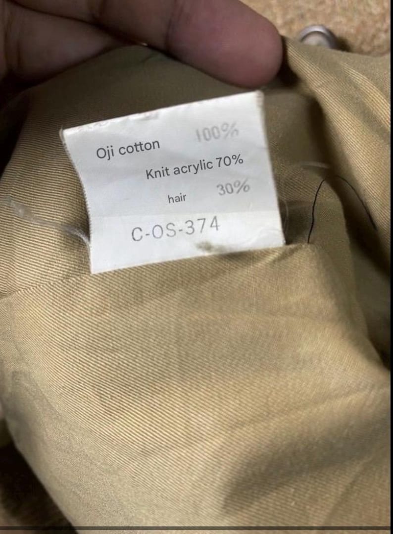 May include: Close-up of a tag with text. The tag reads "Oji cotton 100%, Knit acrylic 70%, hair 30%" and "C-OS-374". The tag is inside a tan fabric.