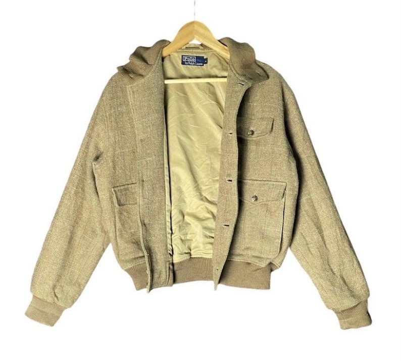 May include: A tan bomber jacket with a hood, two front pockets, and button closures. The jacket has a light brown lining and ribbed cuffs and hem. The Polo by Ralph Lauren logo is visible on the label.