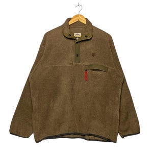 Puede incluir: Un jersey de forro polar marrón con cuello abotonado y un pequeño bolsillo en el pecho. El jersey tiene un acento verde oscuro en el cuello y el bolsillo, con un tirador de cremallera rojo. Un logotipo de huella de pata es visible en el pecho.