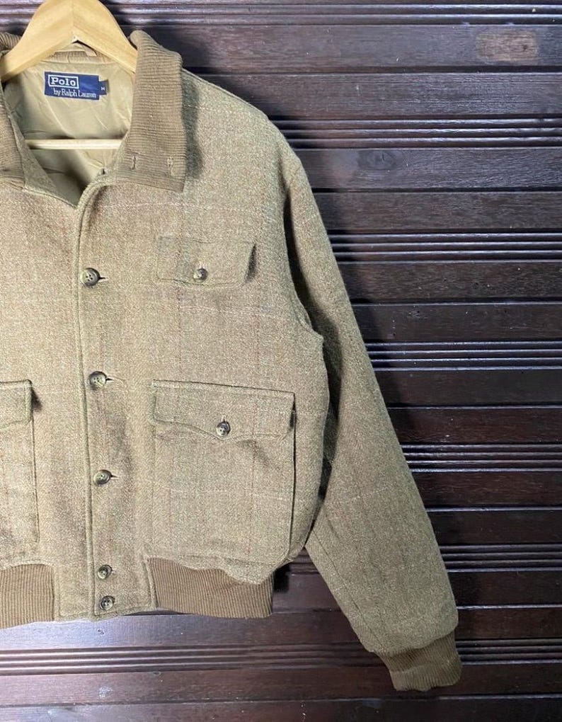 May include: A tan Polo by Ralph Lauren jacket with a corduroy collar and four front pockets. The jacket has a button-up closure and ribbed cuffs and hem. The jacket is hanging on a wooden hanger.