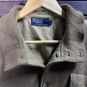 May include: A close-up of a brown Polo by Ralph Lauren jacket. The jacket has a ribbed collar and a button-down front. The Polo label is visible on the inside of the jacket. The jacket is hanging on a wooden hanger.