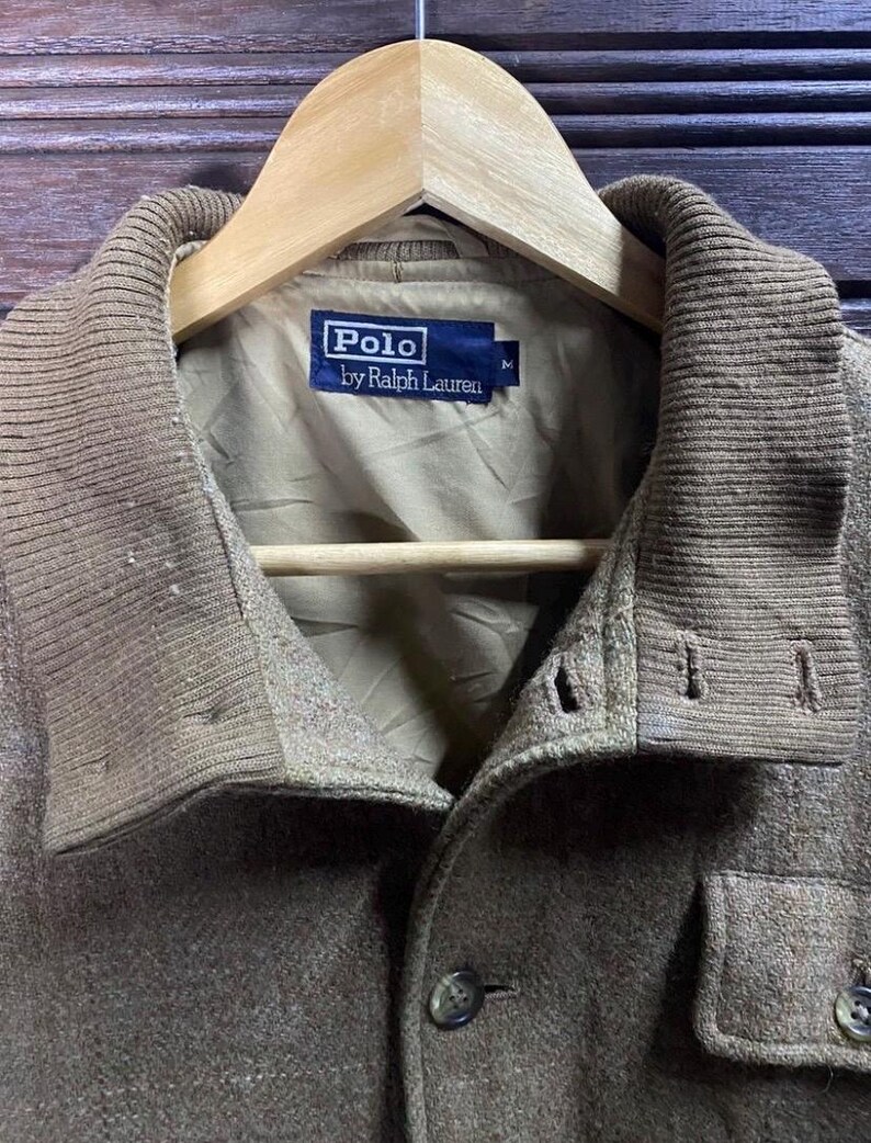 May include: A close-up of a brown Polo by Ralph Lauren jacket. The jacket has a ribbed collar and a button-down front. The Polo label is visible on the inside of the jacket. The jacket is hanging on a wooden hanger.