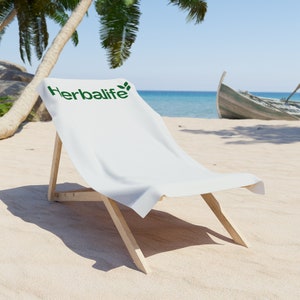 Pode incluir: Uma toalha de praia branca com o logotipo da Herbalife impresso nela, colocada em uma cadeira de praia de madeira em uma praia de areia.