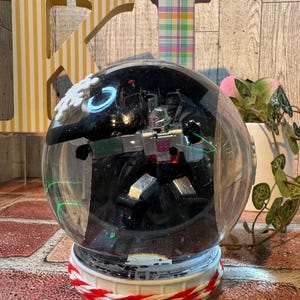 May include: A clear snow globe featuring a silver and purple robot figure. The globe sits on a white base with red and white rope detailing. The background includes wooden letters and a plant.