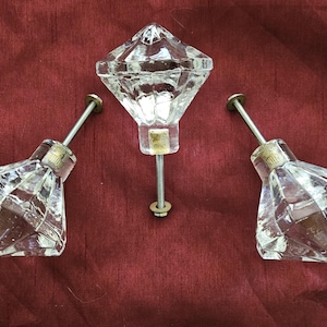 May include: Three clear, faceted glass cabinet knobs with metal stems and hardware. The knobs are diamond-shaped and sit against a deep red background. The central knob is upright, while the other two are angled.