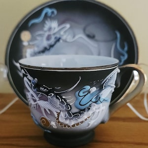 May include: A black porcelain teacup and matching saucer set featuring a raised, three-dimensional white and light blue dragon design. The cup has a gold-toned handle and rim, while the saucer displays a coordinating dragon motif in a matte finish.
