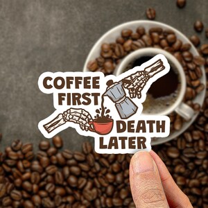 May include: A die-cut sticker featuring two skeletal hands, one pouring coffee from a grey moka pot into a red cup. The text reads COFFEE FIRST DEATH LATER in brown block letters. The design is set against a background of scattered coffee beans.