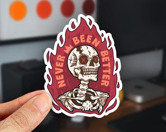 May include: A die-cut sticker featuring a white skull with red X-shaped eyes, set against a dark red flame background. The text 'NEVER BEEN BETTER' is arched around the skull in a light orange font, creating a bold and graphic visual design.