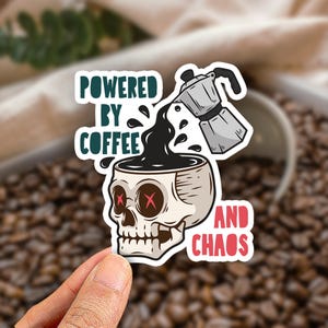 May include: A die-cut sticker featuring a beige human skull with red X-shaped eyes, being filled with black coffee from a silver moka pot. The text reads "POWERED BY COFFEE" in dark green and "AND CHAOS" in red, arranged around the central graphic.