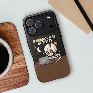 Puede incluir: Una funda de smartphone negra con un gráfico de un esqueleto sentado ante un portátil y el texto "PARANORMAL ACTIVITY". El diseño incluye una luna creciente, una pequeña corona y una taza roja sobre una superficie de escritorio marrón.