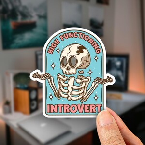 May include: A die-cut sticker featuring a cartoon skeleton with a shrug gesture against a light blue background. The text reads HIGH FUNCTIONING at the top and INTROVERT at the bottom in pink letters. Small white stars are scattered around the skeleton.