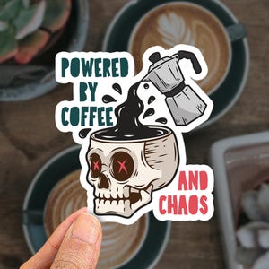 May include: A die-cut sticker featuring a skull with red X-shaped eyes serving as a cup. A silver moka pot pours black coffee into the skull. The text reads POWERED BY COFFEE in dark green and AND CHAOS in red, arranged around the central illustration.