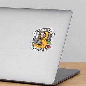 May include: A sticker featuring a skeleton wearing a yellow hoodie, reading a book while sitting in front of a tombstone. A red mug sits nearby. The text LET ME READ IN PEACE is arched around the illustration in bold black capital letters.