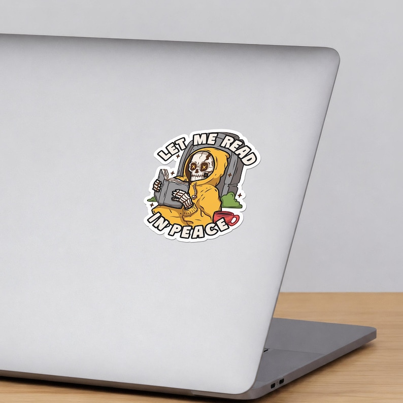 May include: A sticker featuring a skeleton wearing a yellow hoodie, reading a book while sitting in front of a tombstone. A red mug sits nearby. The text LET ME READ IN PEACE is arched around the illustration in bold black capital letters.