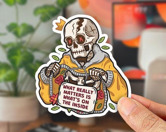 May include: A die-cut sticker featuring a skeleton wearing a yellow hoodie and a small crown. The skeleton holds a white rectangular sign with the text "WHAT REALLY MATTERS IS WHAT'S ON THE INSIDE" surrounded by small red flowers and green leaves.