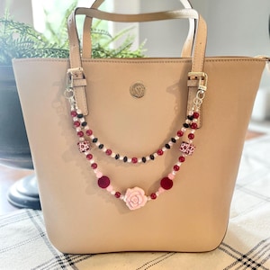 May include: A tan tote bag with a decorative beaded strap. The strap features red, black, and pink beads, including a pink rose accent. The bag has a gold emblem and handles.
