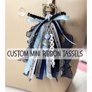 May include: A custom mini ribbon tassel with a silver metal ring. The tassel features various ribbons in blue, navy, and silver, with a beaded charm spelling "BANDOM". The text "CUSTOM MINI RIBBON TASSELS" is displayed on the image.