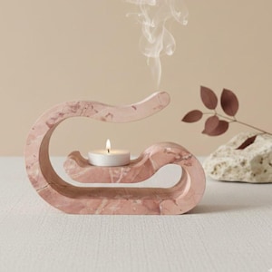 Può includere: Un portacandele in marmo rosa con una candela accesa. Il supporto ha un design curvo unico con una piccola piattaforma per tenere la candela. Volute di fumo si alzano dalla candela, suggerendo che sia profumata. Un oggetto decorativo per l'arredamento della casa.