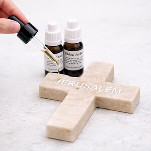 May include: A stone cross with "JERUSALEM" inscribed in white. Two small brown bottles with white lids and labels are behind the cross. A dropper is dispensing a clear liquid onto the cross. The bottles are labelled "Cedar" and "Biblical Amber".