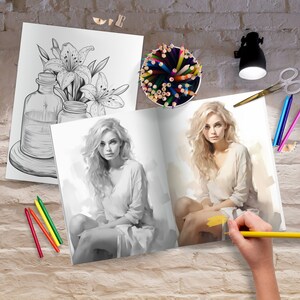 May include: An open coloring book with a black and white illustration of a woman on the left page and a colored illustration of a woman on the right page. The book is on a brick wall surface with colored pencils, a pencil sharpener, and a lamp.