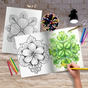 May include: Two open coloring books with floral designs, one colored with green hues. A hand is coloring with a yellow pencil. Colored pencils, scissors, and a small lamp are on a brick surface.