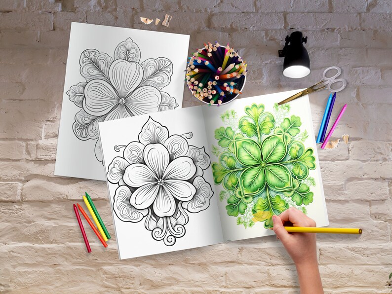 May include: Two open coloring books with floral designs, one colored with green hues. A hand is coloring with a yellow pencil. Colored pencils, scissors, and a small lamp are on a brick surface.