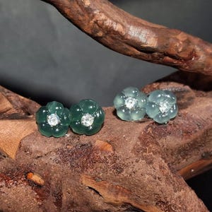 May include: Two pairs of flower-shaped stud earrings. One pair is a deep green color, and the other is a light green color. Each earring features a small, round, clear stone in the center. The earrings are displayed on a piece of dark brown wood.