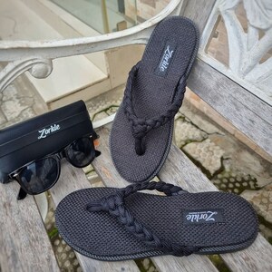 Könnte beinhalten: Ein Paar schwarze Flip-Flops mit geflochtenem Riemen-Design. Die Flip-Flops haben ein schwarzes, gewebtes Obermaterial und eine schwarze Sohle. Der Markenname "Zorkie" ist auf dem Riemen jedes Flip-Flops aufgedruckt.