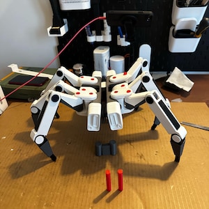 May include: A multi-legged robotic spider model featuring a white and dark grey frame with small red circular accents on the joints. The mechanical creature has eight articulated limbs and is positioned with two separate red cylindrical components.