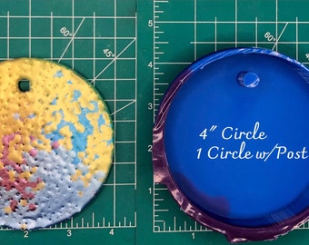 Op de afbeelding: Een ronde harsmal van 10 cm met een pen om op te hangen. De mal is blauw en heeft een paarse rand. De mal ligt op een groene snijmat met een rasterpatroon.