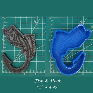 Puede incluir: Un molde de silicona azul de un pez con un anzuelo. El molde está sobre una estera de corte verde con marcas blancas. El molde mide aproximadamente 7,6 cm por 10,8 cm. El texto "Fish & Hook ~3" x 4.25" está debajo del molde.