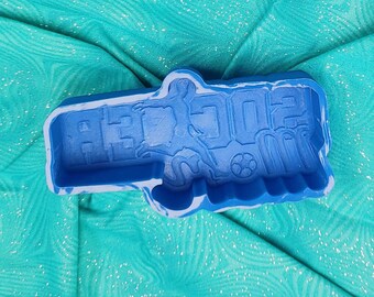 May include: A blue mold with the word "SOCCER" and a soccer player silhouette. The mold also features a soccer ball. The mold is on a teal fabric background.