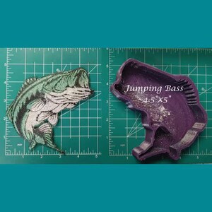 May include: A purple silicone mold of a jumping bass fish. The mold is 4.5 inches by 5 inches. The mold is on a green grid background. There is a green and white embroidered patch of a bass fish on the left side of the image.