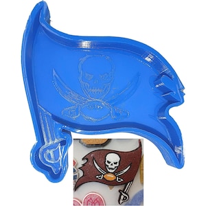 Puede incluir: Cortador de galletas de plástico azul en forma de bandera pirata con un diseño de calavera y huesos cruzados. El cortador se muestra junto a una galleta marrón y roja decorada con el mismo diseño.