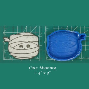 May include: A white and blue mummy-shaped silicone mold for resin crafts. The mold is approximately 4 inches by 3 inches. The mold is on a green cutting mat.