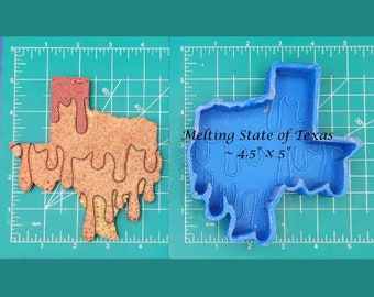Puede incluir: Un molde de silicona azul con forma del estado de Texas con bordes que gotean. El molde está sobre una estera de corte verde con marcas blancas. El texto "Melting State of Texas ~ 4.5" x 5" está impreso en la estera.
