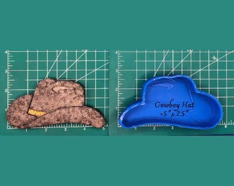 Puede incluir: Se muestran dos objetos con forma de sombrero de vaquero. Uno es de color marrón texturizado con un acento amarillo, el otro es azul liso con el texto "Cowboy Hat ~5" x 2.5"" escrito. Ambos están sobre una cuadrícula verde. El sombrero azul mide aproximadamente 12,7 cm x 6,35 cm.