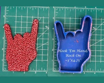 May include: Blue silicone mold of a hand making the rock on hand gesture. The mold is on a green cutting mat with a ruler. The mold is labeled "Hook 'Em Hand/Rock On ~3"X4.75""