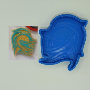 Puede incluir: Molde de plástico azul para hacer una decoración de galleta o pastel con forma de casco de caballero. El casco está delineado en oro y tiene un diseño turquesa.