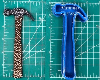 Puede incluir: Un molde de silicona azul en forma de martillo, que mide 6,35 cm por 14 cm, sobre una estera de corte verde con una regla. El molde es para hacer un martillo texturizado.