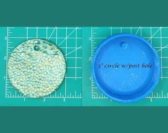 Op de afbeelding: Een ronde harsmal van 7,6 cm met een postgat. De mal is blauw en heeft een getextureerd oppervlak. De mal wordt getoond naast een afgewerkt harsstuk met een vergelijkbaar ontwerp.
