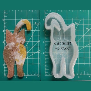 May include: A clear silicone mold for making resin crafts. The mold is shaped like a cat's rear end and is approximately 2.5 inches by 5 inches. The mold is on a green cutting mat with white markings.