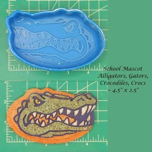 May include: Blue plastic alligator cookie cutter with a detailed design. The cutter is 4.5 inches by 2.5 inches.  The cutter is shown next to a ruler for size reference.  The cutter is also shown next to a finished cookie with a green and brown alligator design.