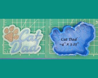 May include: Blue silicone mold for resin casting. The mold is shaped like the words "Cat Dad" with a paw print. The mold measures approximately 4 inches by 3.25 inches.