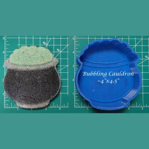 May include: A blue silicone mould of a cauldron with the text "Bubbling Cauldron ~4" x 4.5" ". The mould is on a green cutting mat with a black and green cauldron filled with green glitter.
