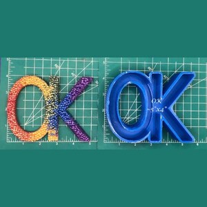 May include: A blue plastic mold for making resin letters. The mold is shaped like the letters "OK" and is approximately 5 inches by 4 inches. The mold is sitting on a green cutting mat.
