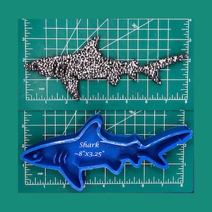 May include: Two shark-shaped items are displayed. The top one is a black and white patterned shark. The bottom one is a blue shark-shaped cookie cutter with the text "Shark ~8"x3.25"" on it. Both are on a green surface.