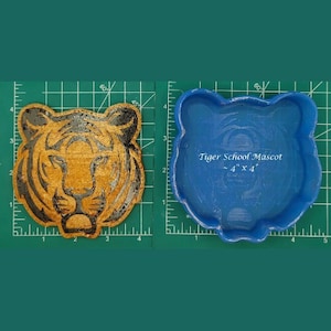 May include: Two tiger-shaped items. One is a detailed, golden-brown tiger head, and the other is a blue, empty mold. The blue mold has the text "Tiger School Mascot" and dimensions of 4" x 4". Both are on a green grid.