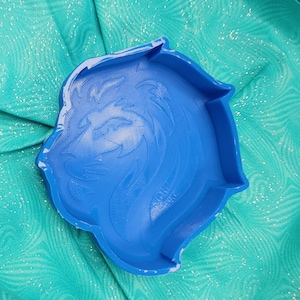 Könnte beinhalten: Eine leuchtend blaue, löwenförmige Schale mit einem detaillierten, geprägten Design eines Löwenkopfes. Die Kunststoffschale liegt auf einem türkisfarbenen Stoffhintergrund mit einem dezenten, schimmernden Muster. Das Gesicht des Löwen ist im Profil.