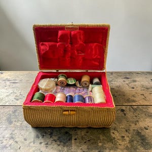 May include: A vintage sewing box with a woven gold exterior and a red velvet lining. Inside are spools of thread in various colours, buttons, and sewing accessories. The box is rectangular and has a wooden clasp.